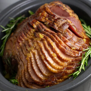 A side view of a spiral cut crock pot ham still in the slow cooker surrounded by fresh rosemary sprigs.