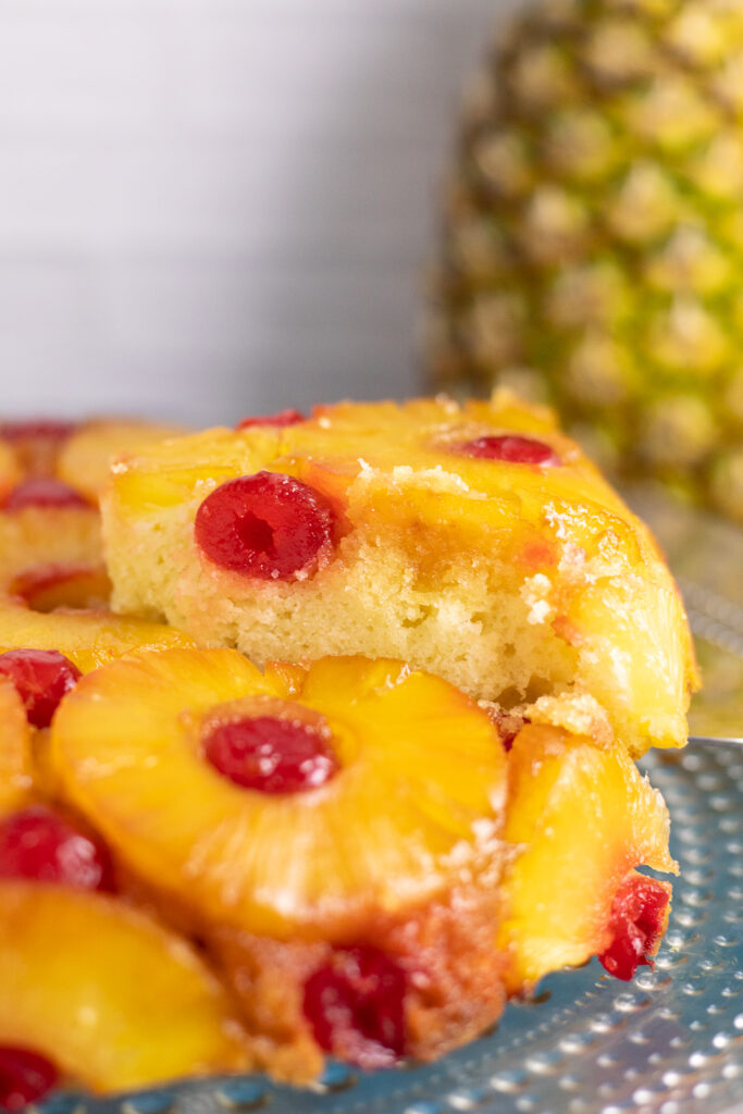 A side view of a slice being lifted with a cake server from a pineapple upside down cake on a glass cake stand with a full pineapple behind.