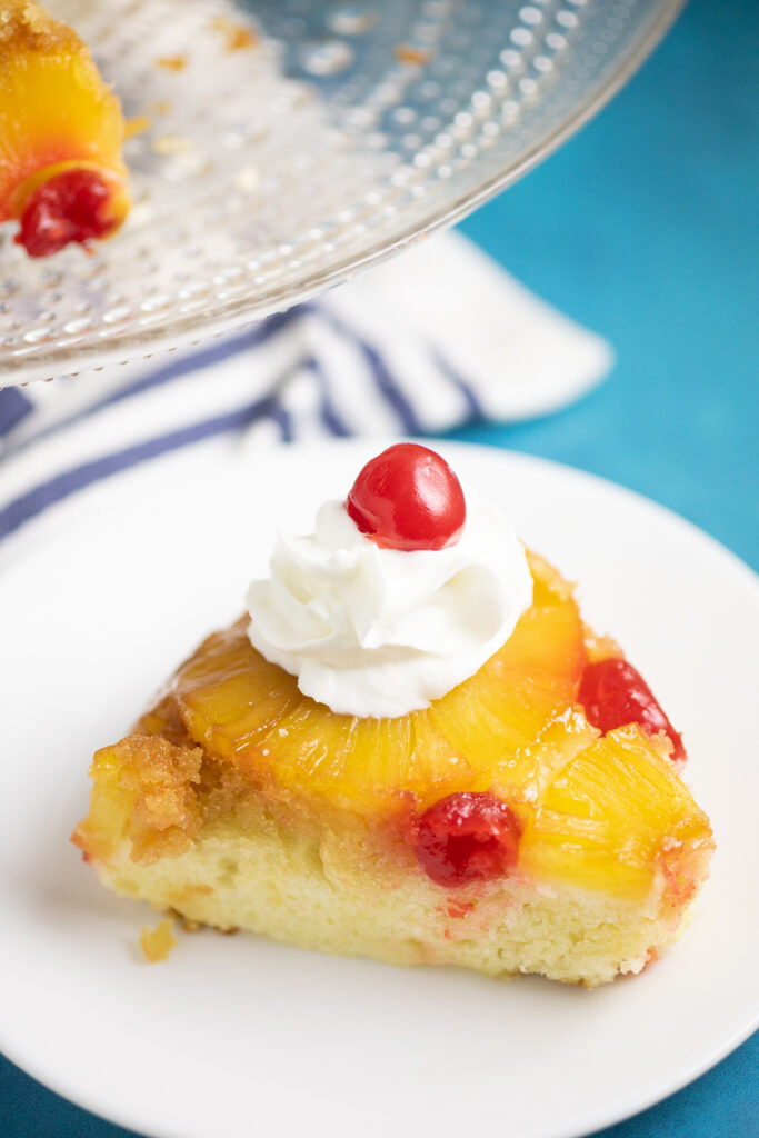 A slice of pineapple upside down cake topped with a dollop of whipped cream and a maraschino cherry on a small plate with a linen napkin and the full cake on a glass cake stand behind.