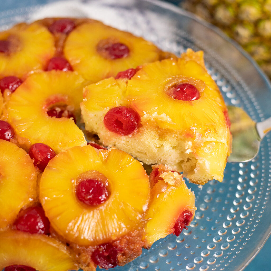 A slice being lifted from a pineapple upside down cake on a glass cake stand with a full pineapple behind.