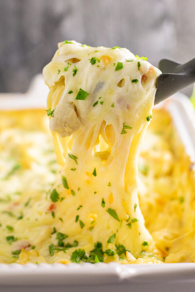 A silicone spoon scooping out a large cheesy portion of Rotel chicken spaghetti topped with fresh cilantro from a large casserole dish with gooey melty cheese stretching from the dish.