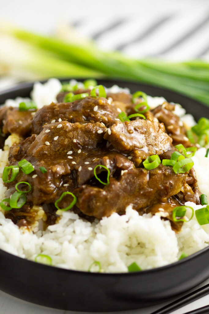 Slow Cooker Mongolian Beef in a bowl over white rice, topped with sliced green onions and sesame seeds with a linen napkin and green onions behind.