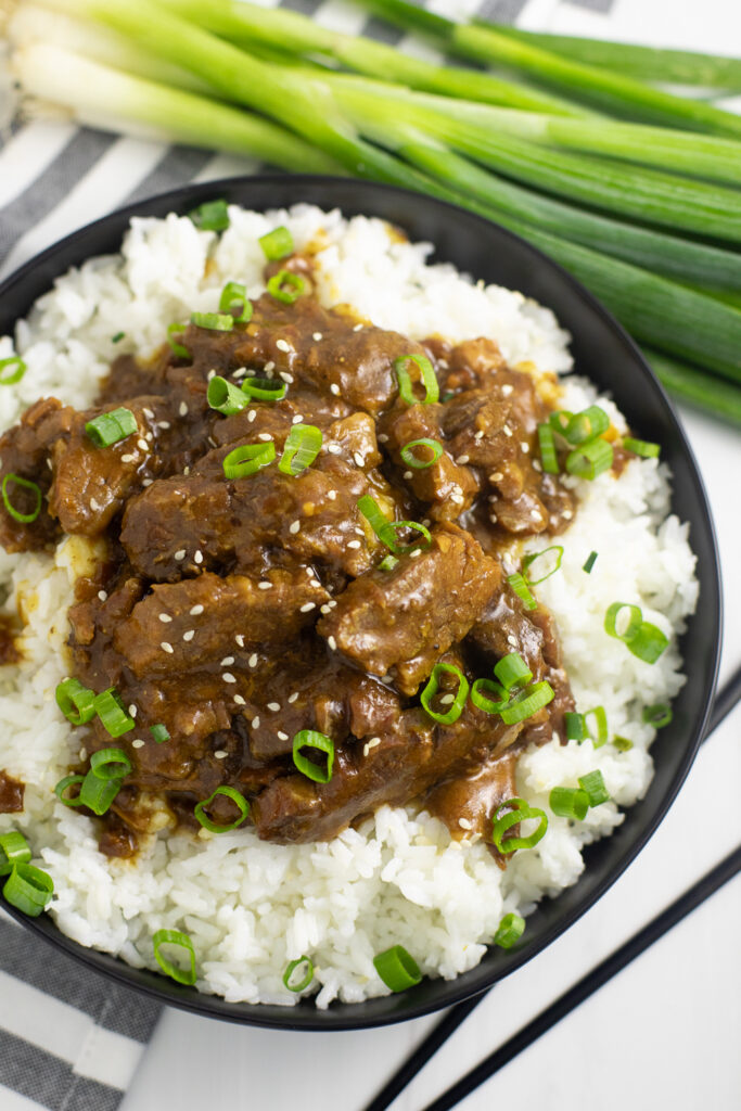 Overhead view of slow cooker Mongolian beef served over white rice in a black bowl next to green onions and linen napkin.