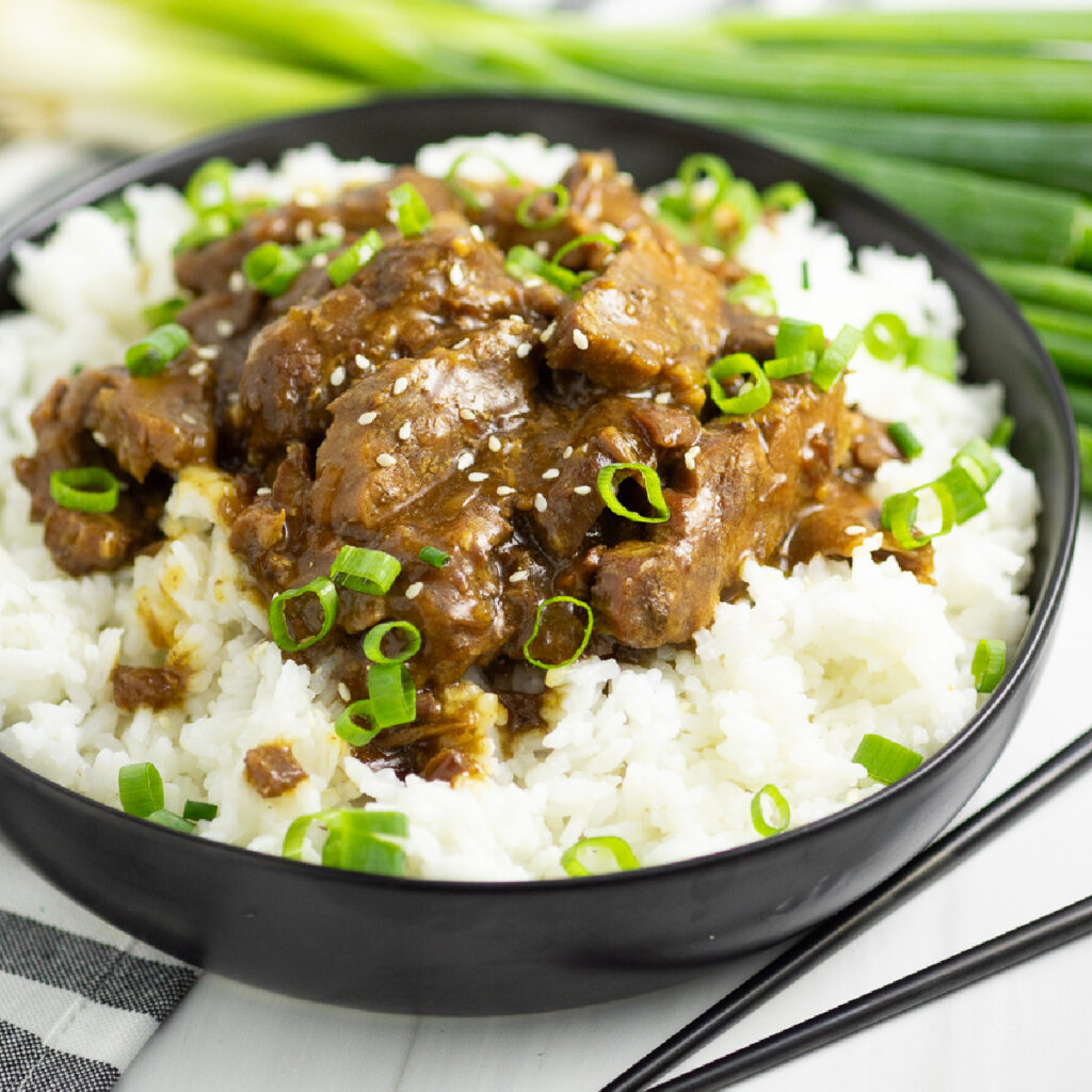 Slow Cooker Mongolian Beef in a bowl over white rice, topped with sliced green onions and sesame seeds next to chopsticks and a linen napkin with green onions behind.