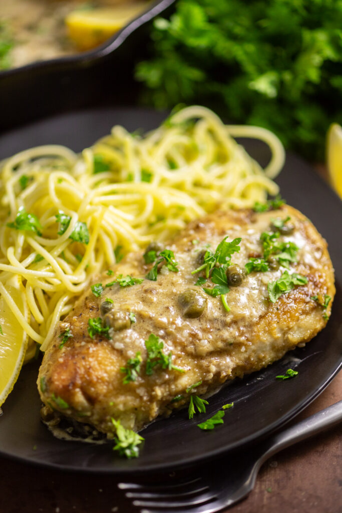 Chicken scallopini on a small black plate next to pasta topped with freshly chopped parsley.