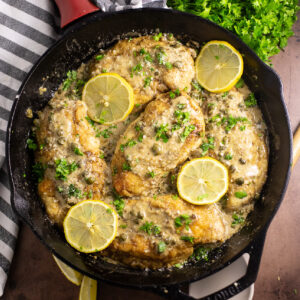 Overhead view of chicken scallopini in a cast iron skillet with lemon slices topped with freshly chopped parsley. The skillet is next to a linen napkin and a fresh bunch of parsley on a rustic background.