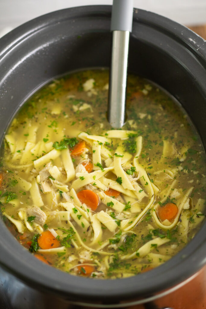 A slow cooker filled with chicken noodle soup topped with freshly chopped parsley with a ladle in the middle.