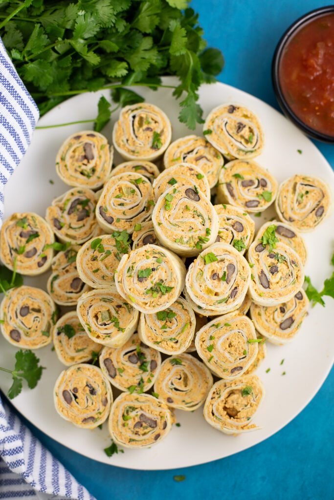 Mexican pinwheels stacked on a large white plate topped with freshly chopped cilantro next to a fresh bunch of cilantro, a small bowl of salsa, and a linen napkin.