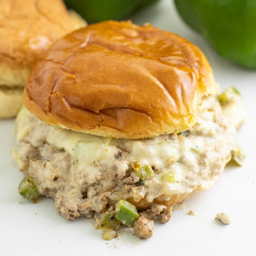 Overhead view of a Philly cheesesteak sloppy joe topped with melted provolone and a toasted bun with another sandwich and two green bell peppers behind it.