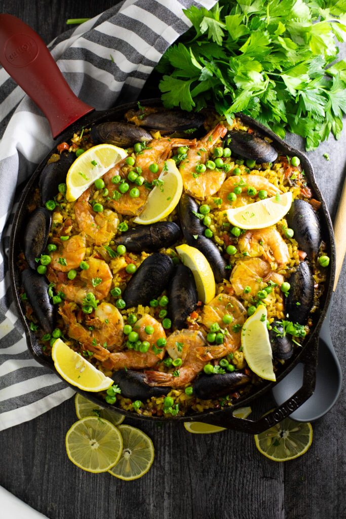 Overhead view of Spanish paella topped with mussels, shrimp, peas, and lemon wedges in a cast iron skillet next to a fresh bunch of parsley, linen napkin, and lemon slices.