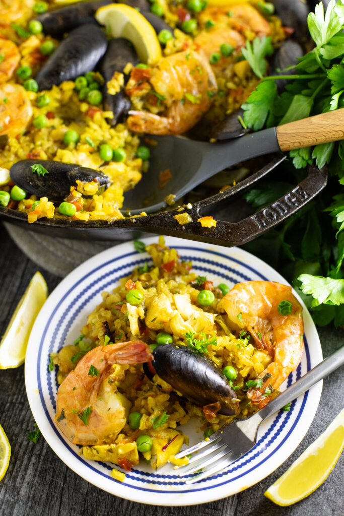 A scoop of Spanish paella on a small plate with the remaining skillet of paella behind with a wooden spoon in it.