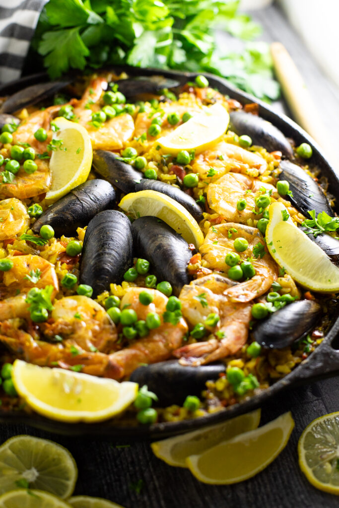Close up of a Spanish seafood paella topped with peas, mussels, shrimp, and lemon wedges in a cast iron skillet with a bunch of parsley behind.