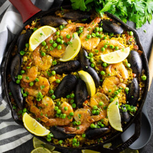 Overhead view of Spanish paella topped with mussels, shrimp, peas, and lemon wedges in a cast iron skillet next to a fresh bunch of parsley, linen napkin, and lemon slices.