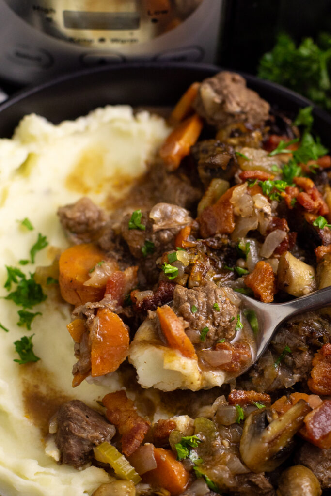 A spoon scooping up a bite of slow cooker beef bourguignon and mashed potatoes topped with freshly chopped parsley.
