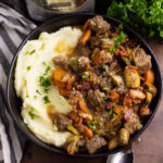 Overhead view of slow cooker beef bourguignon in a shallow bowl next to mashed potatoes topped with freshly cut parsley. The bowl is next to a linen napkin and a fresh bunch of parsley.