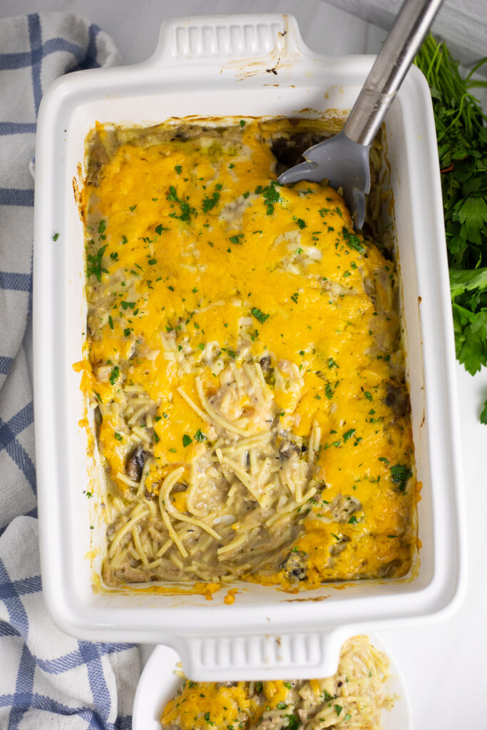 Overhead view of a large ceramic casserole dish filled with tuna tetrazzini with a scoop missing, topped with melted cheese and freshly chopped parsley.