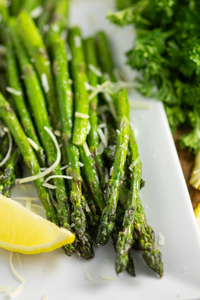 Close up of air fryer asparagus on a rectangle plate with a lemon wedge, next to a bunch of curly parsley.