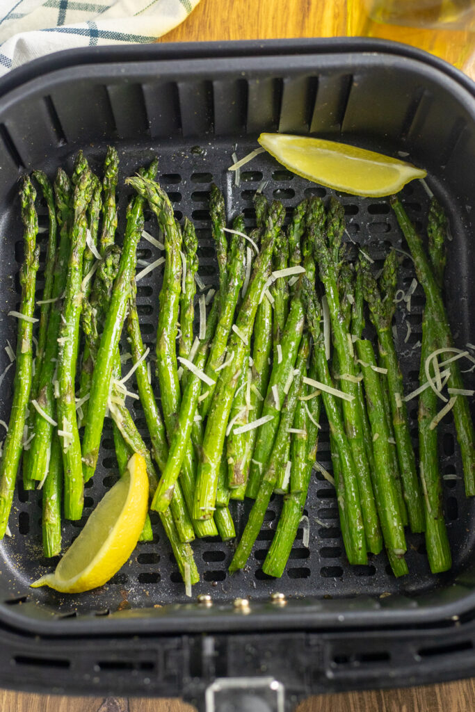 Asparagus in an air fryer basket topped with shredded parmesan and lemon wedges.