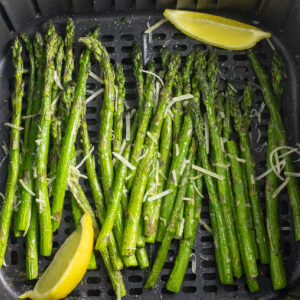 Asparagus in an air fryer basket topped with shredded parmesan and lemon wedges.