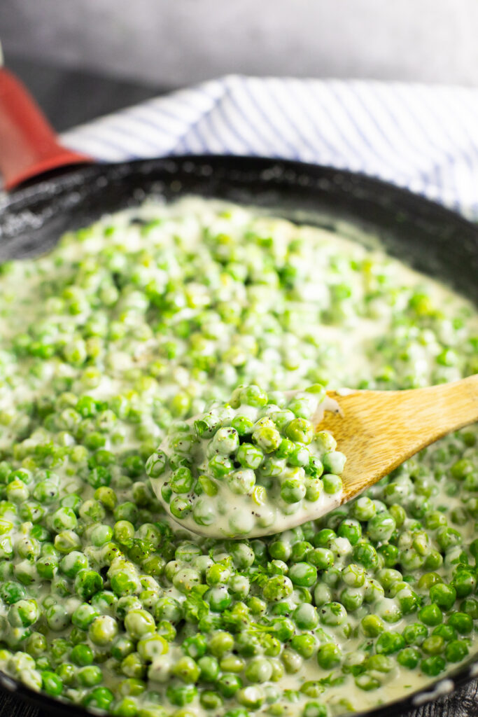 A wooden spoon taking a scoop of creamed peas out of a cast iron skillet.