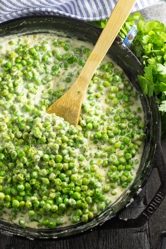 Overhead view of a cast iron skillet filled with creamed peas with a wooden spoon in the center, surrounded by a bunch of fresh parsley and a striped linen napkin.