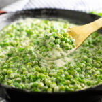 A wooden spoon taking a scoop of creamed peas out of a cast iron skillet.