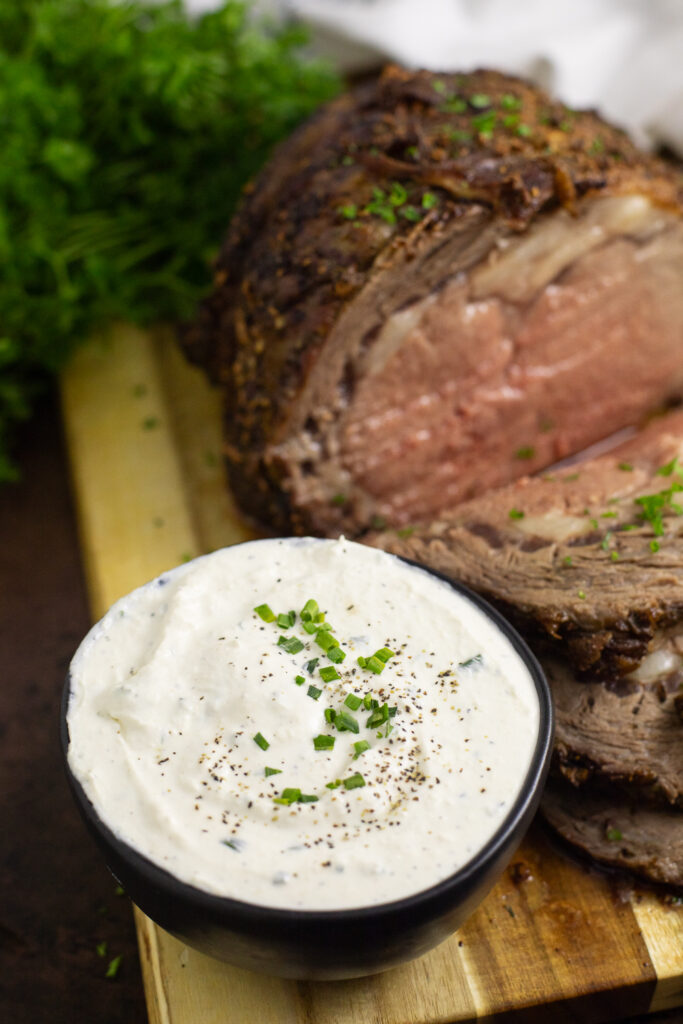 Creamy horseradish sauce in a small black bowl topped with black pepper and fresh sliced chives next to prime rib on a cutting board.