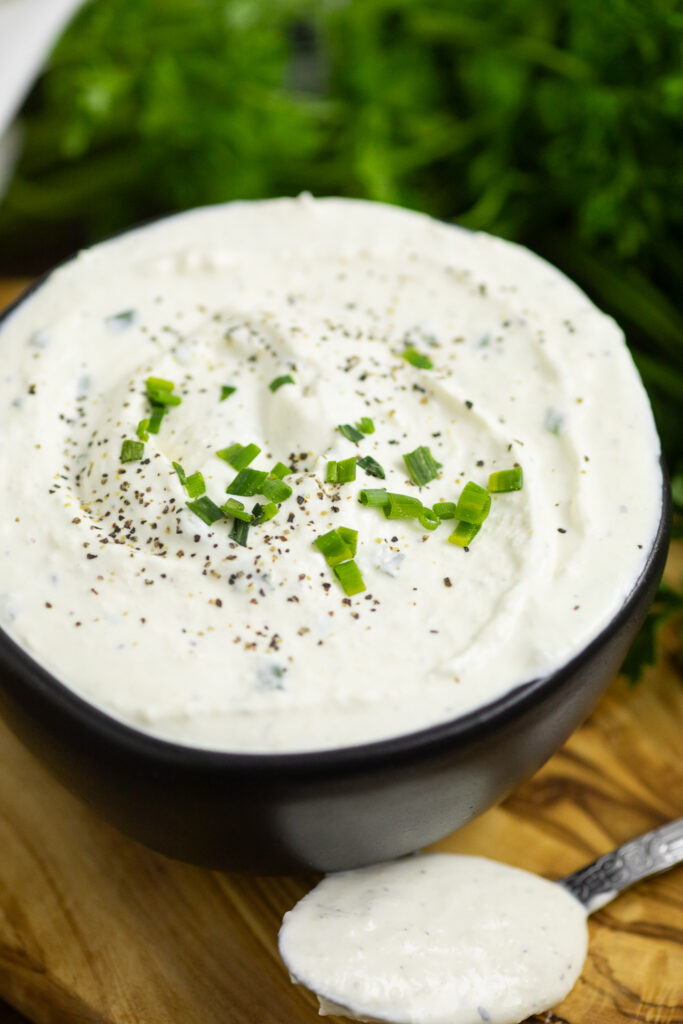 Close up of creamy horseradish sauce in a small black bowl topped with black pepper with fresh sliced chives with a spoon filled with sauce in the front and a bunch of parsley behind, all on a wooden cutting board.
