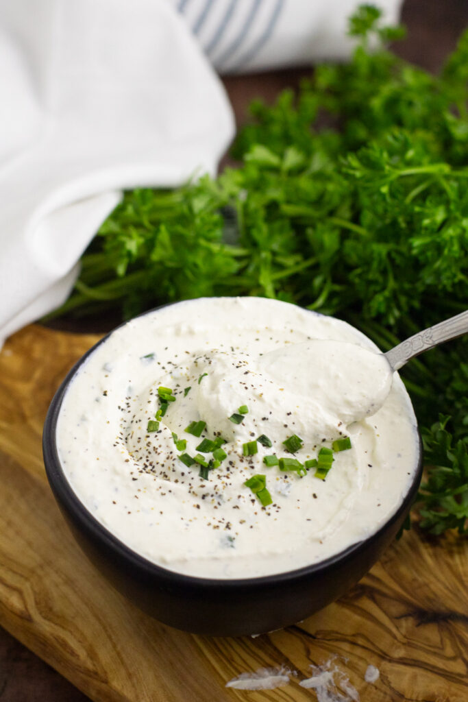 Close up of creamy horseradish sauce in a small black bowl topped with a spoon taking a scoop out and a bunch of parsley behind, all on a wooden cutting board.