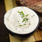 Creamy horseradish sauce in a small black bowl topped with black pepper and fresh sliced chives next to prime rib on a cutting board.