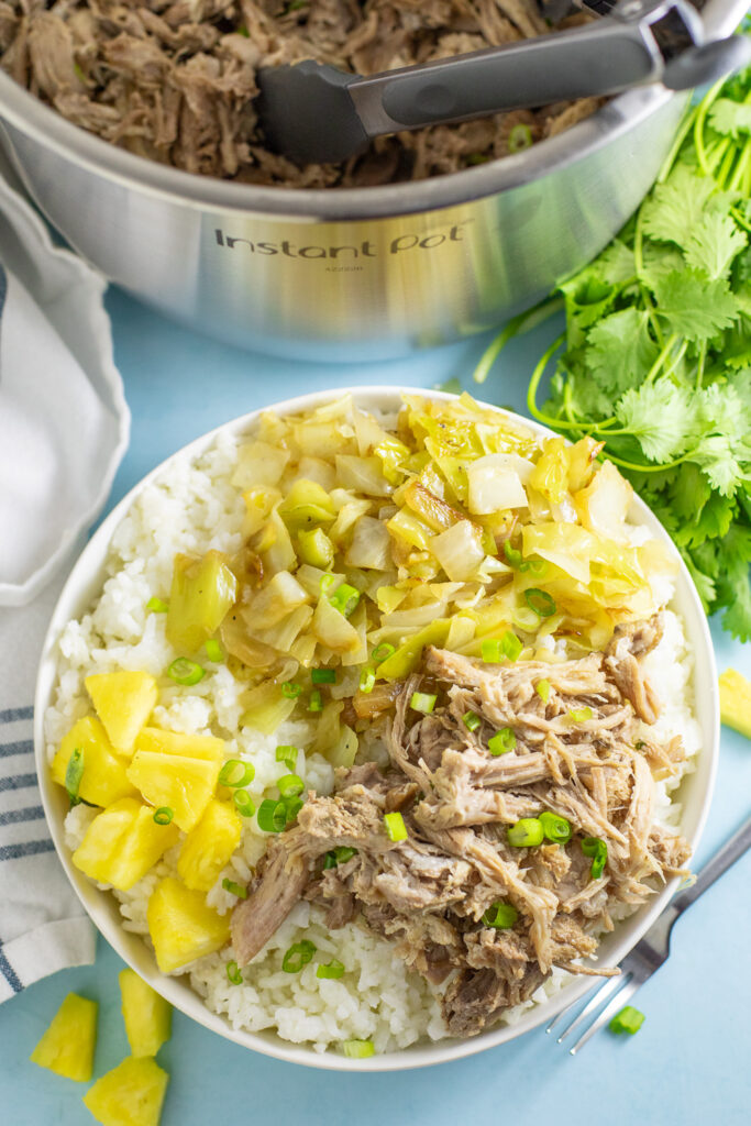 Overhead view of a shallow bowl filled with white rice and topped with instant pot kalua pork, sauteed cabbage, and fresh pineapple, garnished with sliced green onions. Fresh cilantro and a linen napkin are next to the bowl with the instant pot behind.