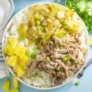 Overhead view of a shallow bowl filled with white rice and topped with instant pot kalua pork, sauteed cabbage, and fresh pineapple, garnished with sliced green onions. Fresh cilantro and a linen napkin are next to the bowl.