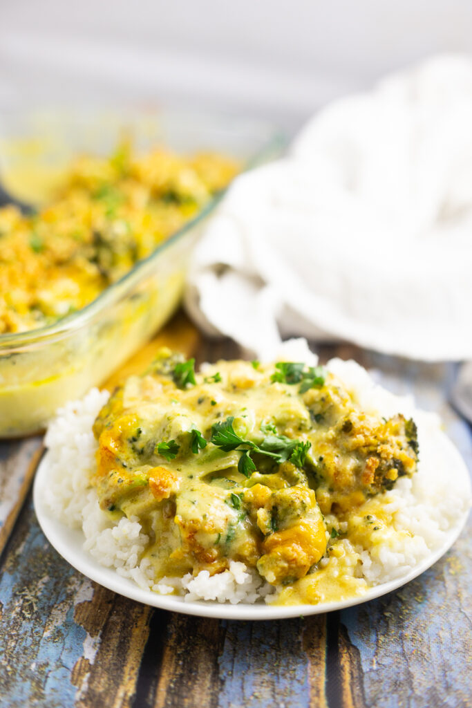 Chicken divan with curry served over a bed of white rice on a small plate with the full glass casserole dish and a linen napkin behind it.