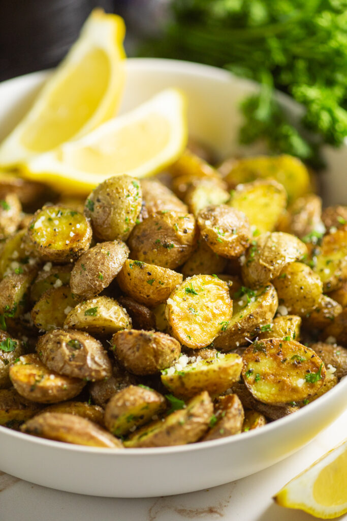 Air fryer baby potatoes in a serving bowl topped with fresh parsley next to two lemon wedges and a fresh bunch of parsley.