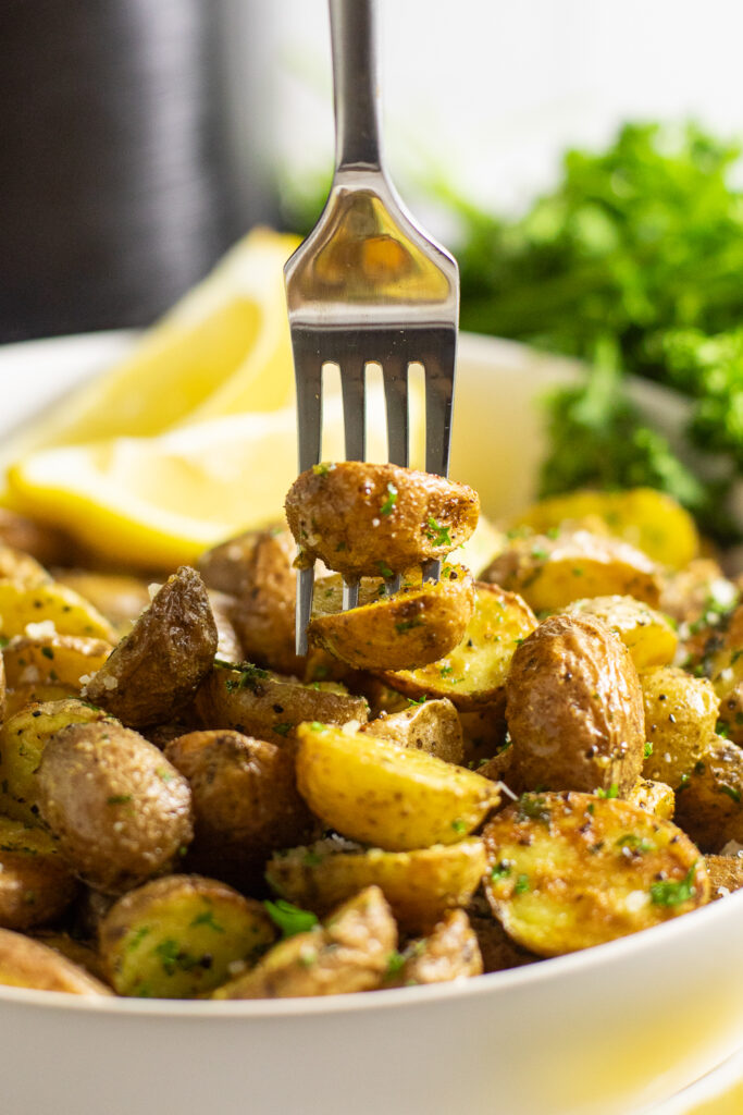 A fork stabbing two baby potatoes over a large serving bowl with more potatoes in it, with lemon wedges and a bundle of fresh parsley in the background.