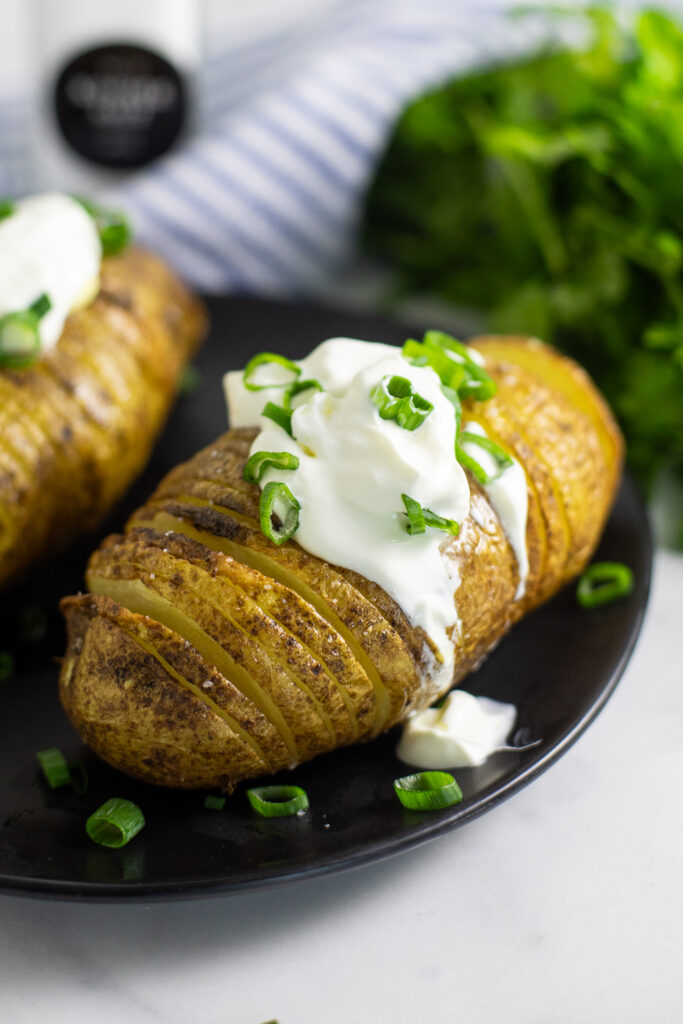 Air fryer hasselback potato topped with sour cream and green onions with a bunch of parsley and a linen napkin behind.
