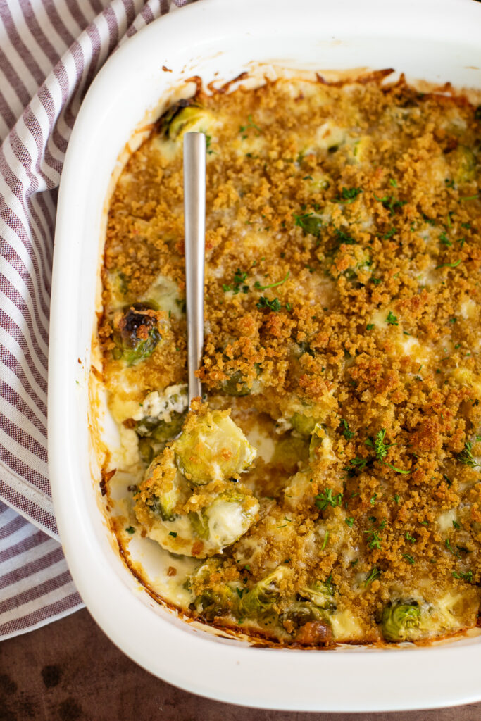 Overhead view of a white casserole dish with brussels sprouts gratin topped with crispy breadcrumb topping and a spoon take a scoop.