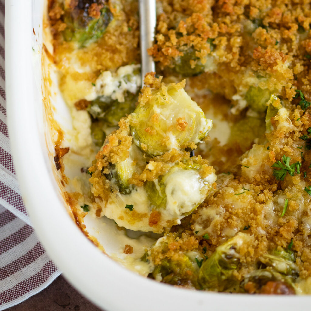 Overhead view of a white casserole dish with brussels sprouts gratin topped with crispy breadcrumb topping and a spoon take a scoop.