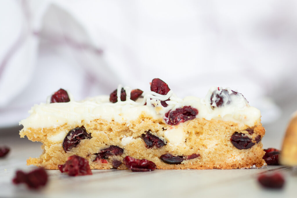 Side view of a cranberry bliss bar on a rustic wood surface, showing the inside of the bar studded with white chocolate chips and cranberries.