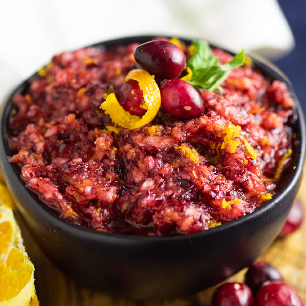 Black matte bowl filled with cranberry relish and topped with fresh cranberries, an orange peel curl, and a mint leaf.