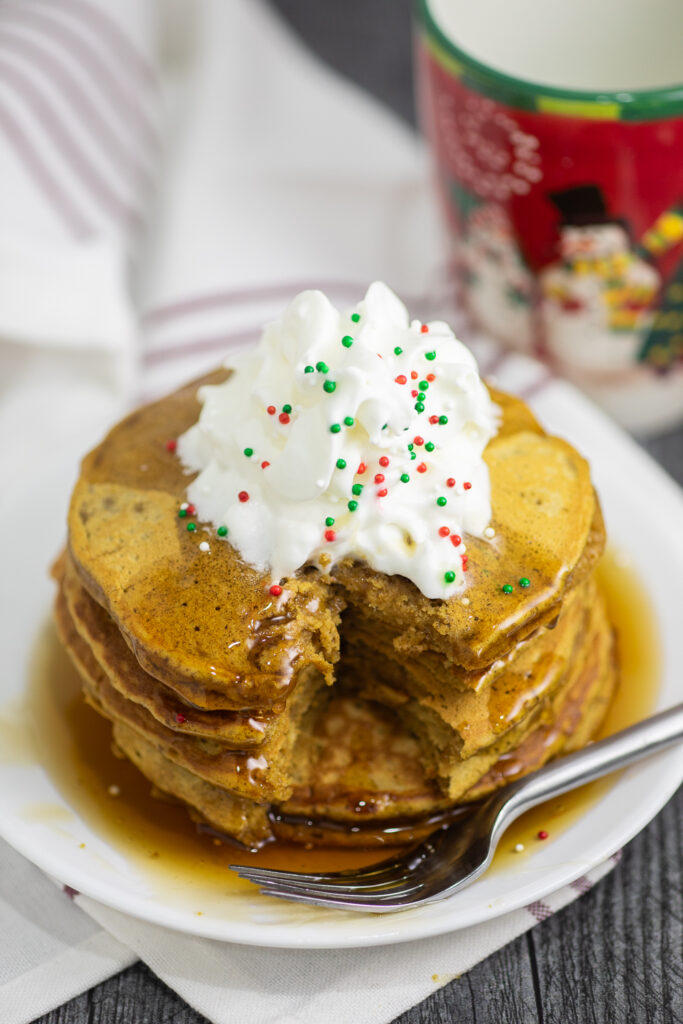 A stack of gingerbread pancakes on a small white plate with a bite taken out next to a fork.