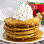A stack of gingerbread pancakes on a small white plate with maple syrup dripping down the stack and whipped cream and sprinkles on top. A Christmas snowman mug and linen napkin are behind.