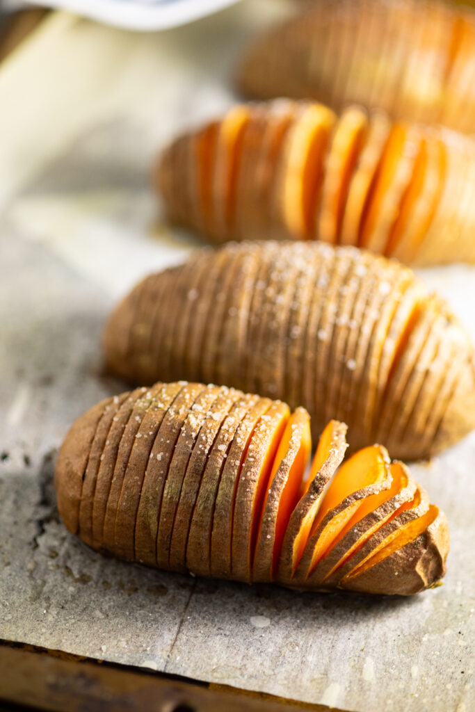 Hasselback sweet potato on a baking sheet topped with flaky sea salt with more sweet potatoes behind.