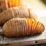 Hasselback sweet potato on a baking sheet topped with flaky sea salt with more sweet potatoes behind.