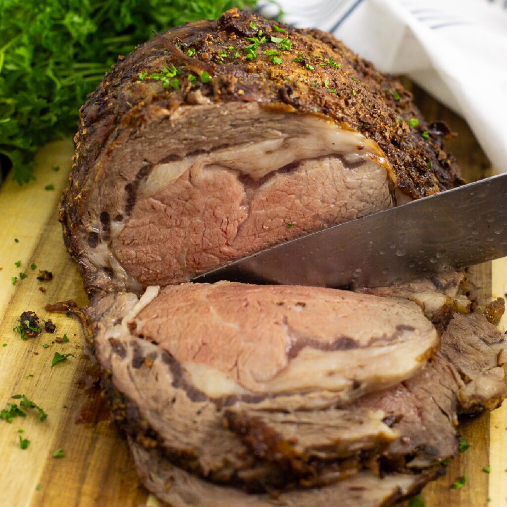 A prime rib roast topped with chopped parsley with a knife cutting a slice off on a wooden cutting board with a bunch of parsley and a linen napkin behind.