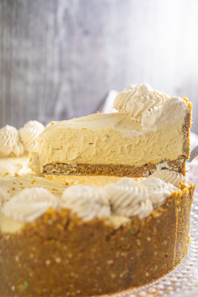 A piece of no bake pumpkin cheesecake being lifted from the full pie on a cake stand with a rustic background.