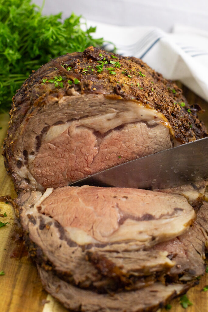 A prime rib roast topped with chopped parsley with a knife cutting a slice off on a wooden cutting board with a bunch of parsley and a linen napkin behind.
