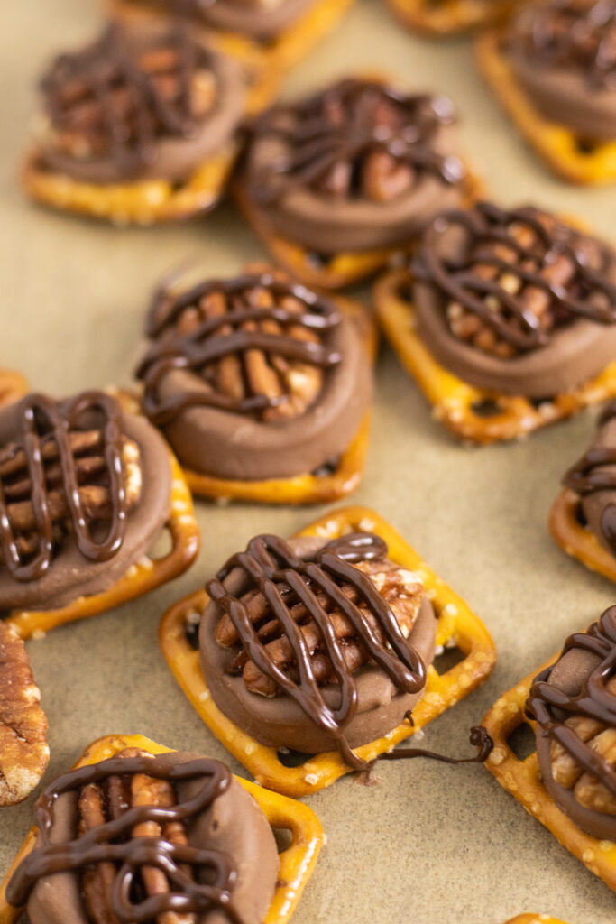 Rolo pretzel turtles drizzled in melted chocolate next to pecans on a baking sheet with parchment paper.