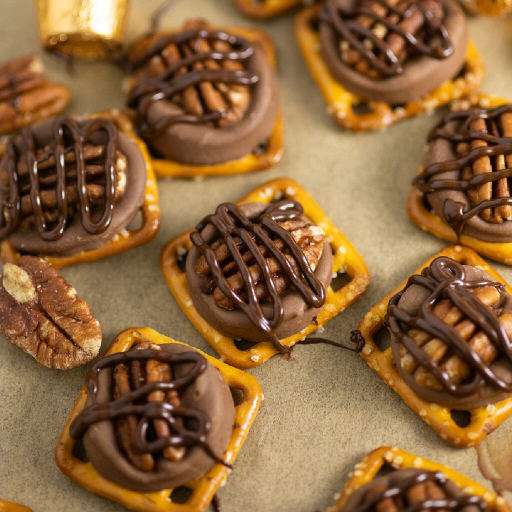 Rolo pretzel turtles drizzled in melted chocolate next to pecans on a baking sheet with parchment paper.