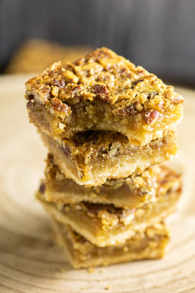 Five southern pecan pie bars stacked on top of each other in a tower on a rustic cutting board. The top bar has a bite taken out.